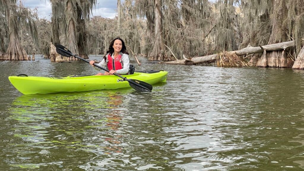 A woman in a red life vest smiles while paddling a bright lime-green kayak through the calm waters of Lake Martin. In the background, weathered cypress trees draped in thick Spanish moss and a large fallen log line the swamp’s shore under a soft winter sky.