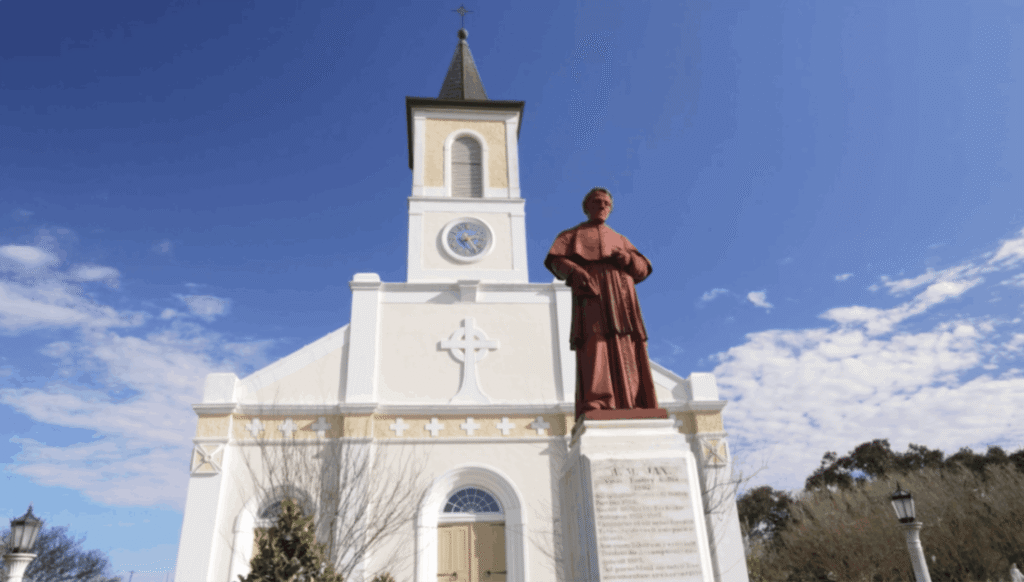 "Exterior view of the white St. Martin de Tours Catholic Church in Saint Martinville, Louisiana, with a large red statue of Father Ange Marie Jan on a stone pedestal in the foreground under a blue sky.