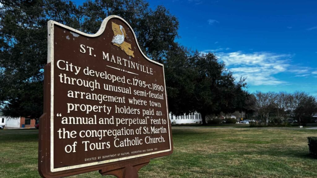 Historical marker for St. Martinville, Louisiana, explaining the city's unusual semi-feudal development from 1795 to 1890 involving property rents paid to St. Martin de Tours Catholic Church. The brown roadside sign stands in a grassy park under a bright blue sky.