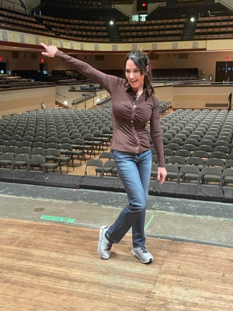 A woman in a brown long-sleeved button-up shirt and blue jeans stands on a wooden stage, pointing one arm toward a large, empty indoor auditorium. The rows of dark gray seats and tiered balcony seating are visible behind her. A green "Shreveport" sticker is on the stage floor at her feet.