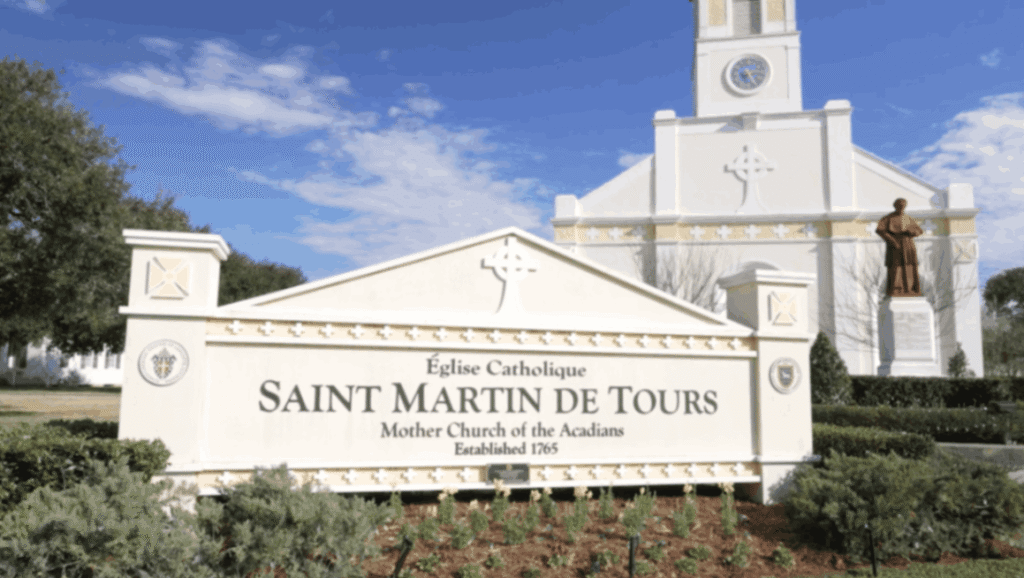 Saint Martin de Tours Catholic Church monument sign in St. Martinville, Louisiana, with the historic church building and statue in the background.