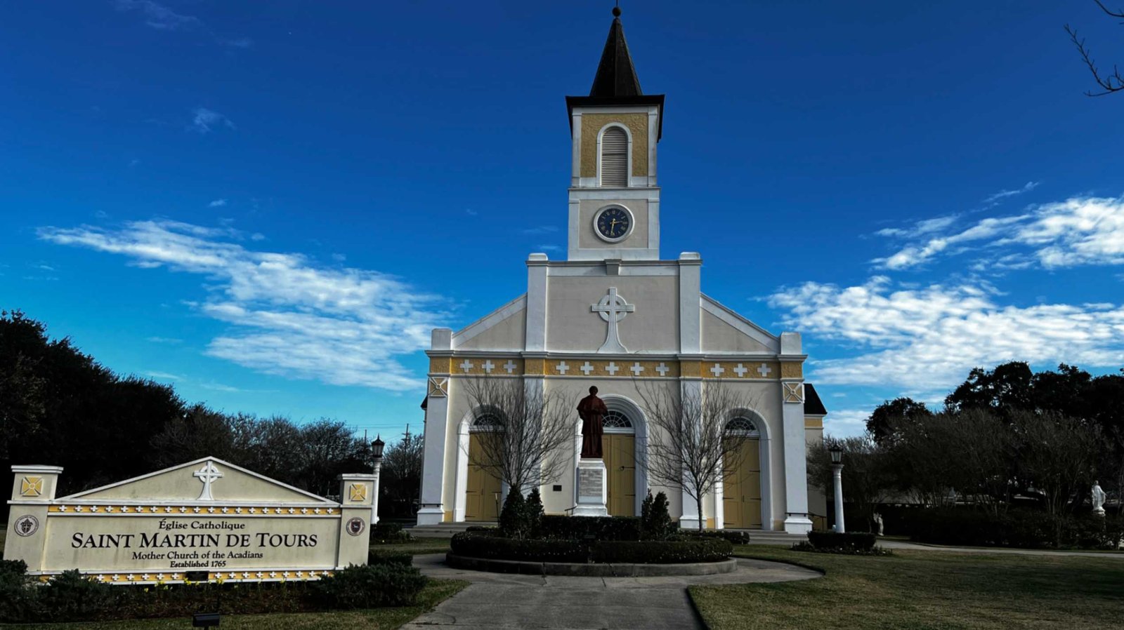 Saint Martin de Tours Catholic Church in St. Martinville, Louisiana, showing the historic cream-colored facade, central bell tower, and entrance with a statue and "Mother Church of the Acadians" signage in the foreground.