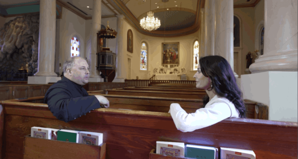 In the sunlit interior of St. Martin de Tours Catholic Church, Father Jason Vidrine, in a black cassock, and Karen LeBlanc, in a white top, sit in wooden pews facing each other for an interview. The background shows the ornate altar, stained glass windows, and a large crystal chandelier hanging from the arched ceiling.