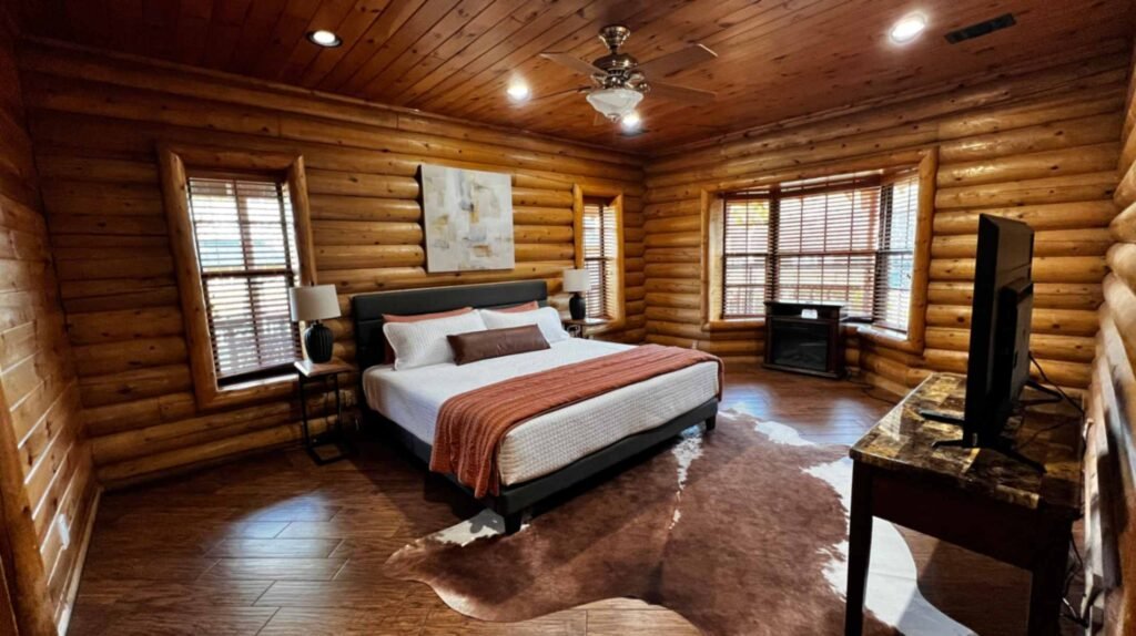 A spacious, rustic master bedroom inside a log cabin at God's Country RV Park. It features log walls and a wood-paneled ceiling, a king-sized bed with a black headboard and orange throw, a large cowhide rug on dark wood floors, and a bay window with a small electric fireplace.