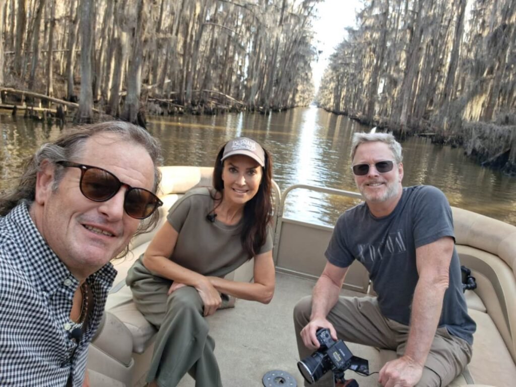 A selfie of three people two men and a woman on a pontoon boat in a narrow waterway on Caddo Lake. One man in the foreground wears sunglasses and a checkered shirt, a woman in the center wears a cap and olive clothing, and another man to the right holds a professional camera. They are surrounded by tall, moss-covered cypress trees lining the water.