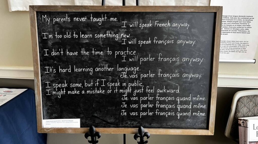 A chalkboard sitting on an easel displays a poem about reclaiming language. The left side lists excuses in English for not speaking French, like 'My parents never taught me' and 'I'm too old to learn.' The right side answers each excuse with a phrase that gradually transitions from English to French, ending with the repeated line, 'Je vas parler français quand même' (I am going to speak French anyway).