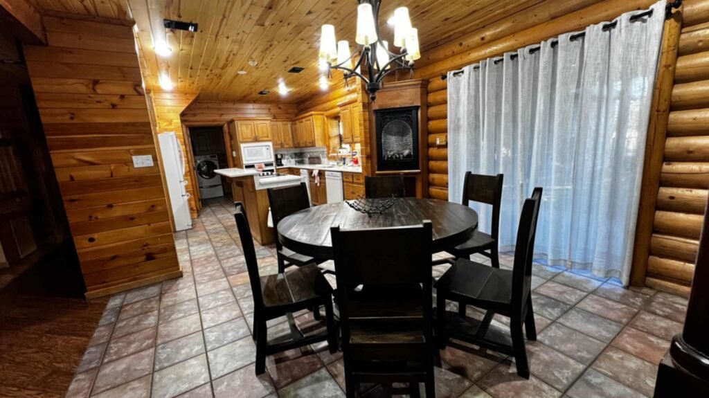The kitchen and dining area of a rustic log cabin at God's Country RV Park. A dark wood round dining table with five matching chairs sits under a wrought-iron chandelier. The space features log walls, tiled floors, and a fully equipped kitchen with wooden cabinets and white appliances in the background.