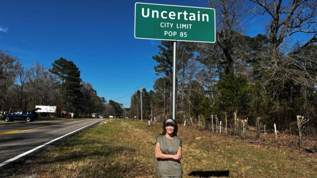 A woman stands smiling beneath a green road sign that reads "Uncertain City Limit, Pop 85." The sign is located along a paved two-lane highway under a clear blue sky, marking the entrance to the small town on the Texas side of Caddo Lake.
