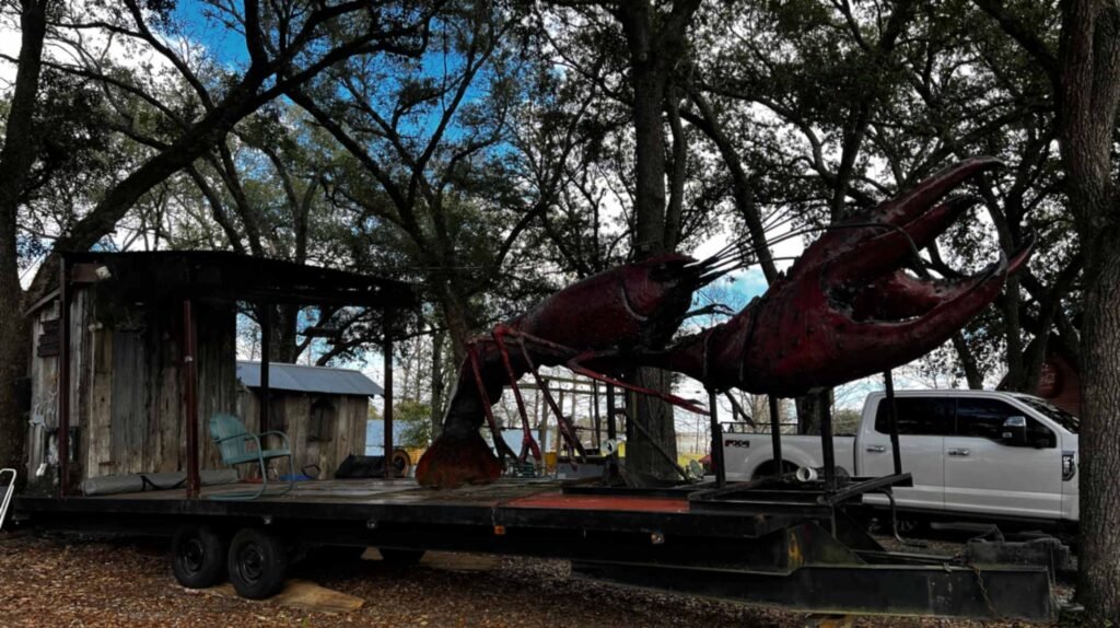 A giant red crawfish sculpture and a small wooden shack on a long flatbed trailer, parked beneath a canopy of large trees at Bayou Cabins in Breaux Bridge, Louisiana.