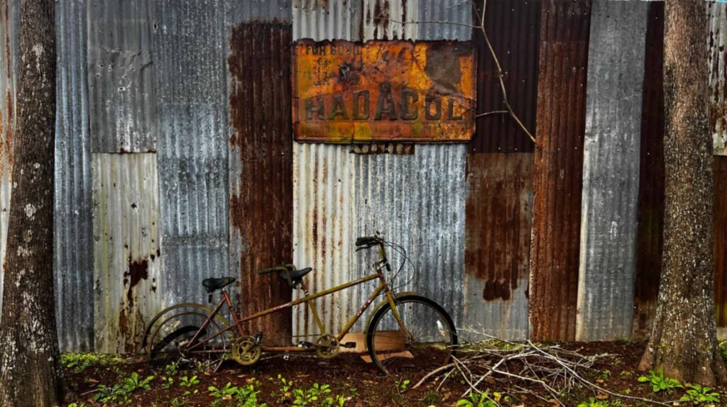 A rusty tandem bicycle leans against a weathered corrugated metal wall with a vintage, rusted "Hadacol" sign at Bayou Cabins in Breaux Bridge.
