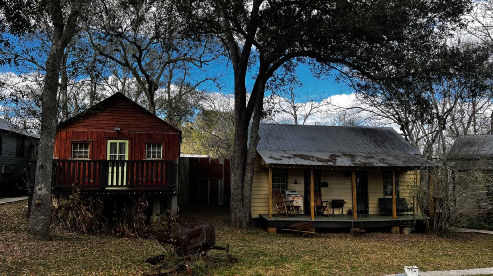 A dark red wooden cabin with a small balcony sits next to a light yellow wooden cabin with a shaded porch, both tucked beneath large, mossy trees at Bayou Cabins.