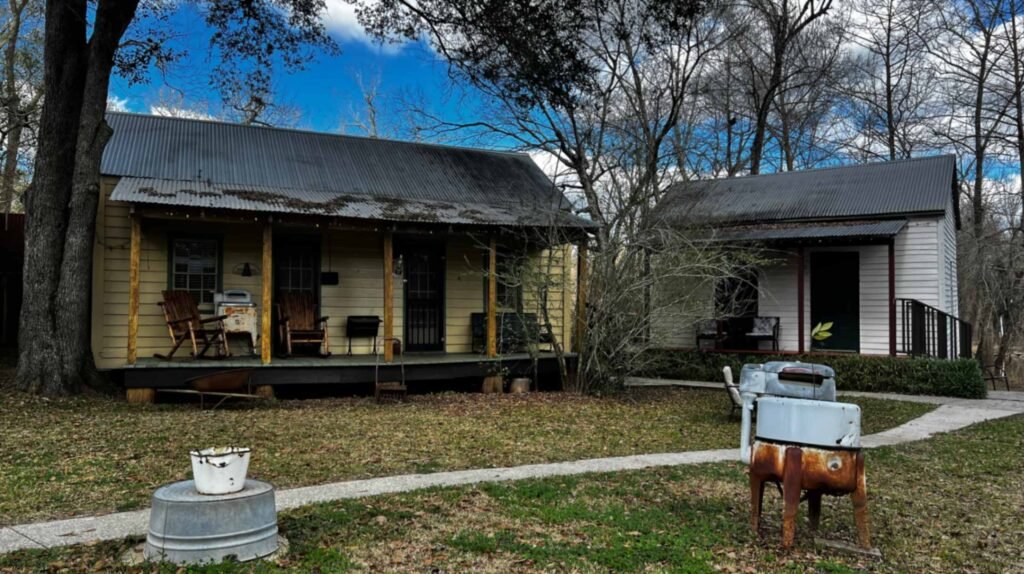 Two small, rustic wooden cabins with corrugated metal roofs and front porches, surrounded by bare trees and featuring vintage metal tubs in the yard.