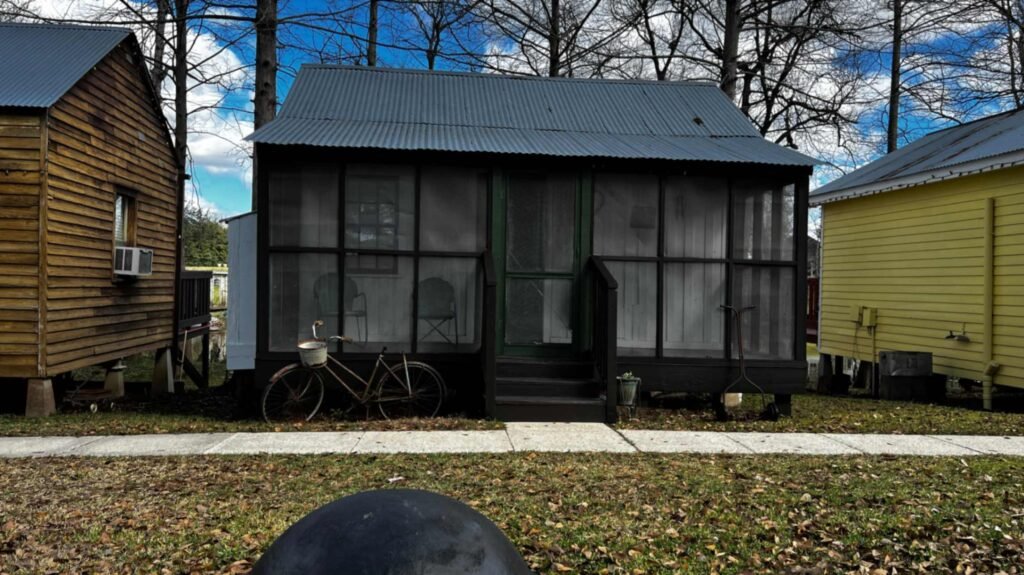 A small dark brown wooden cabin with a screened-in porch and a corrugated metal roof, flanked by a cedar-shingled cabin and a yellow cabin, with a vintage bicycle parked in front at Bayou Cabins in Breaux Bridge.