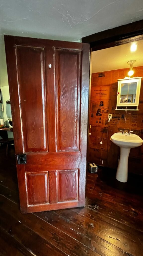 A heavy, dark-stained four-panel wooden door standing open to reveal a rustic bathroom with dark wood floors, a white pedestal sink, and walls covered in vintage newspaper clippings.