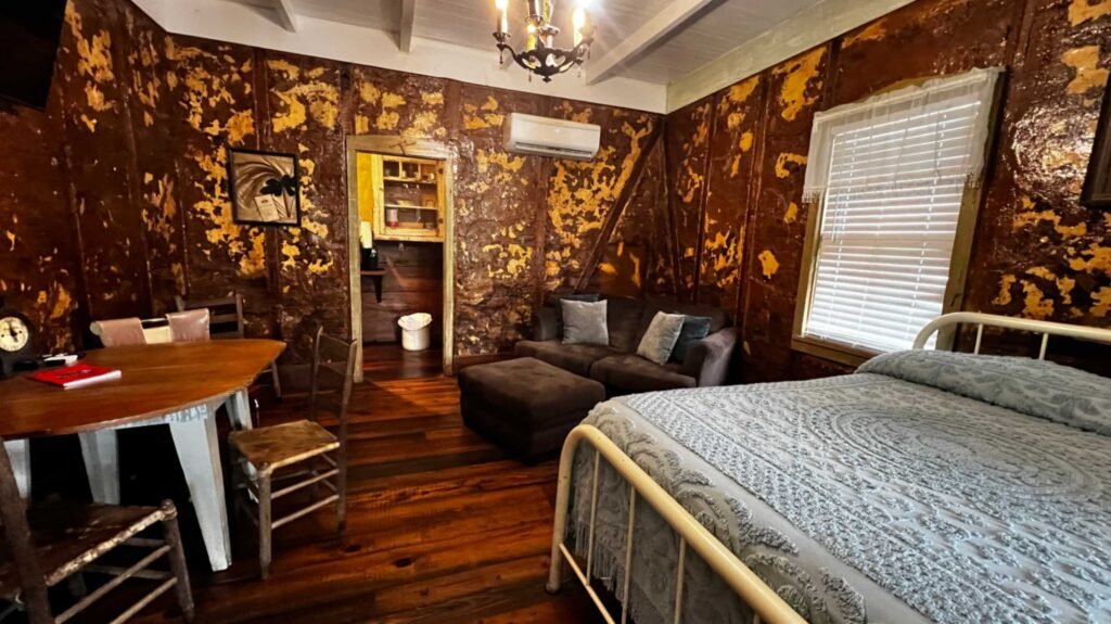 A wide-angle view of a rustic cabin interior with textured brown and amber bousillage walls, wide-plank wood floors, a white metal bed, a small brown sofa, and a vintage dining table.