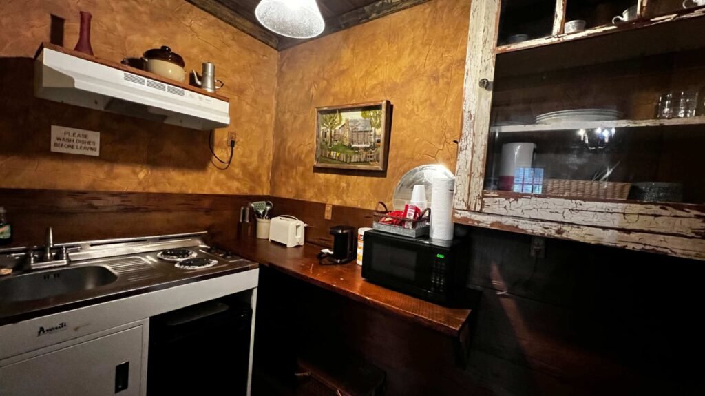 A small, rustic kitchenette with textured amber-colored walls, a dark wood countertop holding a microwave and toaster, and an old white stove unit under a weathered wooden cabinet.