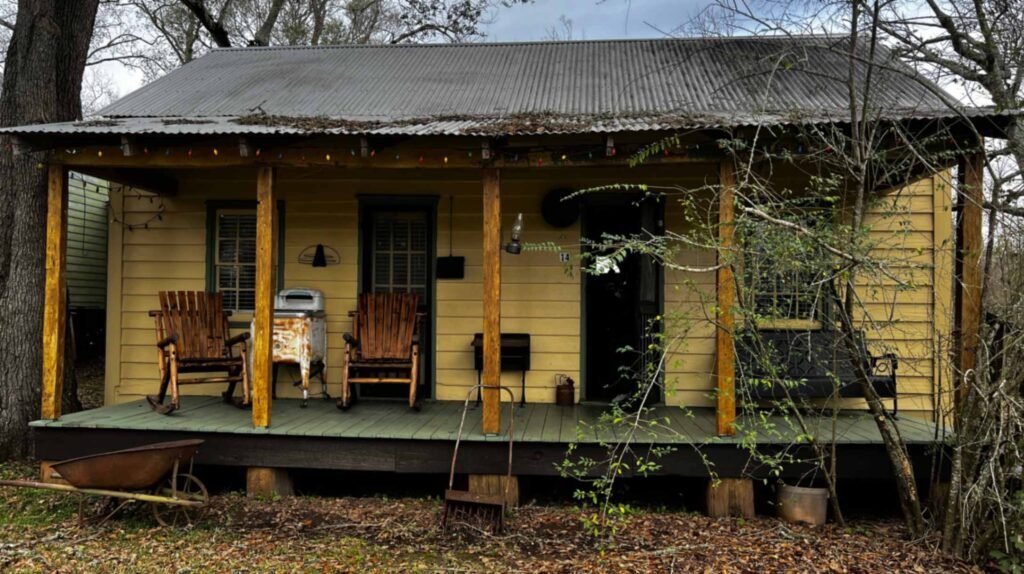 A small yellow wooden cabin with a corrugated metal roof and a green porch featuring two wooden rocking chairs, a vintage metal tub, and a black bench.