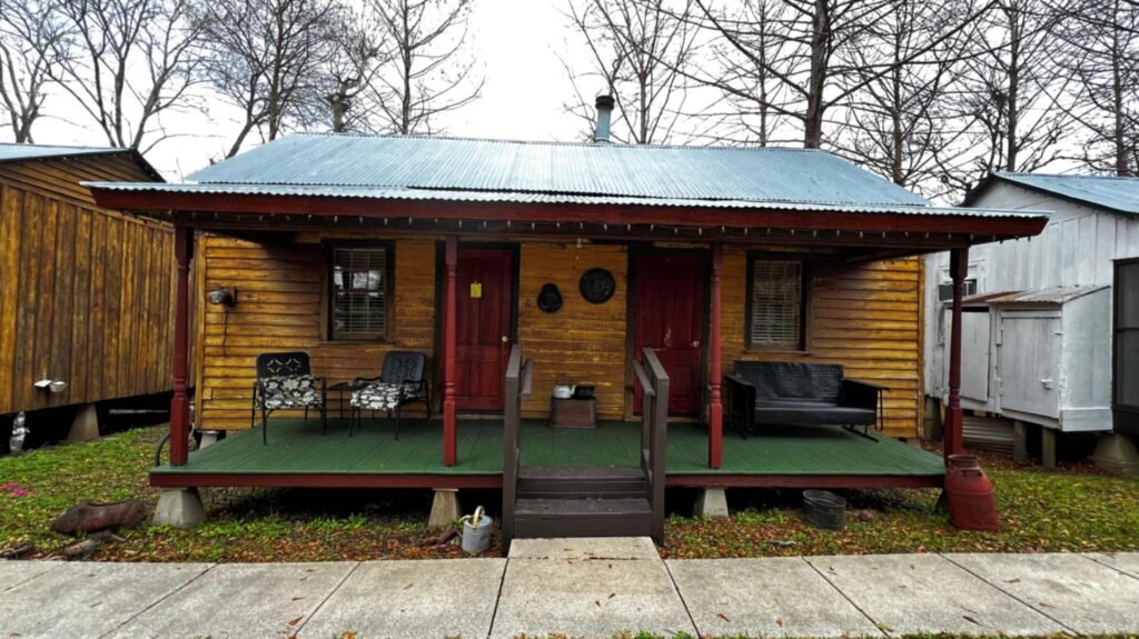 A small, rustic yellow wooden cabin at Bayou Cabins in Breaux Bridge, Louisiana, featuring a green porch with a small set of stairs, two red doors, and a corrugated metal roof, set against a backdrop of bare winter trees.