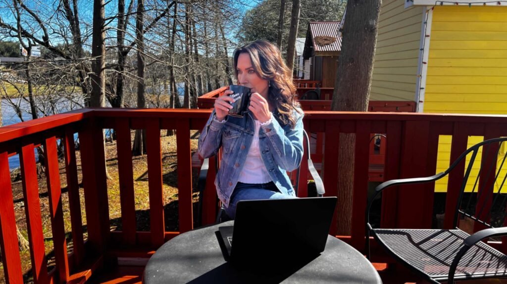 A woman in a denim jacket drinks from a mug while sitting at a small table with a laptop on a red wooden balcony, with Bayou Teche and trees in the background at Bayou Cabins in Breaux Bridge.