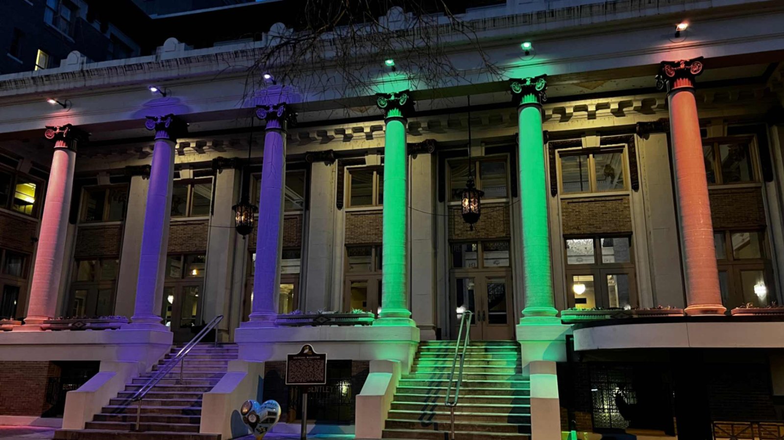 The historic exterior of Hotel Bentley at night, featuring grand white Corinthian columns illuminated with vibrant purple and green lights above wide stone entrance stairs.