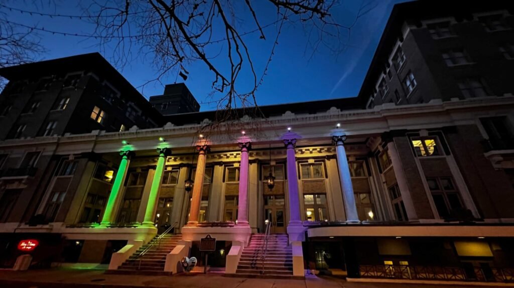 The historic exterior of Hotel Bentley at night, featuring grand white Corinthian columns illuminated with vibrant purple and green lights above wide stone entrance stairs.