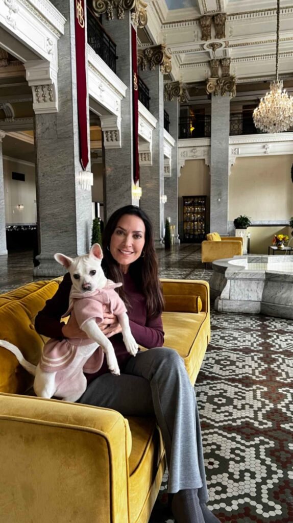 Karen sits on a yellow velvet sofa in the historic grand lobby of the Hotel Bentley in Alexandria, Louisiana, holding Coco Chanel, the hotel's resident dog mascot. In the background are massive marble columns, crystal chandeliers, and the iconic five-color mosaic tile floor.