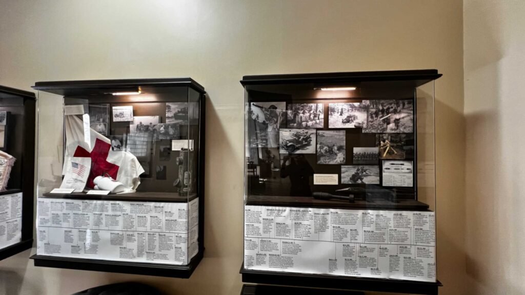 Glass wall-mounted museum display cases at the Hotel Bentley in Alexandria, Louisiana, featuring World War II artifacts, historical photographs of military training, a Red Cross uniform, and a detailed chronological timeline of the Louisiana Maneuvers.