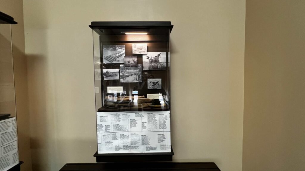 A vertical glass museum display case at the Hotel Bentley in Alexandria, Louisiana, containing black-and-white historical photographs of the Louisiana Maneuvers and a printed chronological timeline of World War II military operations.