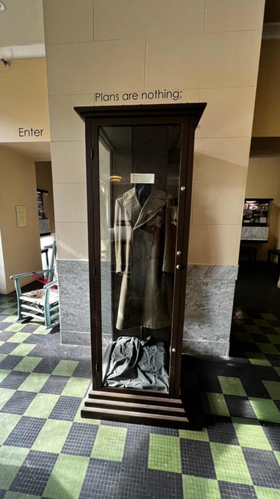 A tall, glass display case at the Hotel Bentley in Alexandria, Louisiana, featuring a vintage World War II military officer's wool overcoat, standing on a checkered floor beneath an wall inscription that reads, "Plans are nothing;".