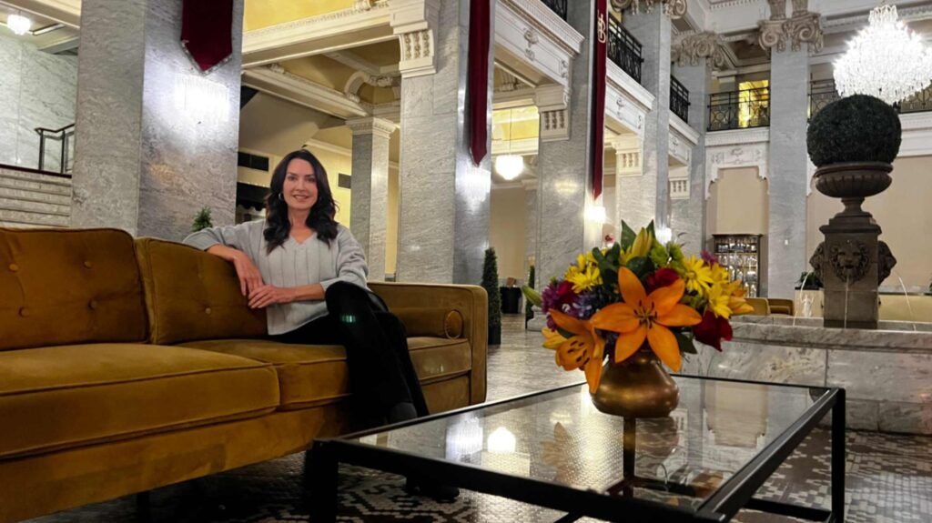 A woman sits on a plush yellow velvet sofa in the grand lobby of the historic Hotel Bentley in Alexandria, Louisiana, featuring a marble fountain, crystal chandeliers, and massive Corinthian columns.