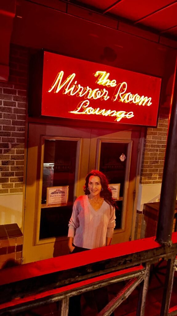 Karen stands in front of the entrance to The Mirror Room Lounge at the Hotel Bentley in Alexandria, Louisiana, beneath a glowing red neon sign. The entrance features dark wood doors and a classic brick facade.
