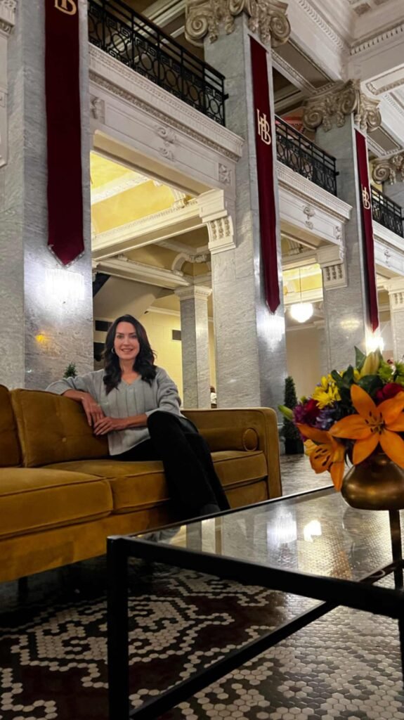 A woman sits on a yellow velvet sofa in the grand lobby of the Hotel Bentley in Alexandria, Louisiana, featuring soaring marble columns, ornate horsehair plaster moldings, and the historic five-color mosaic tile floor.