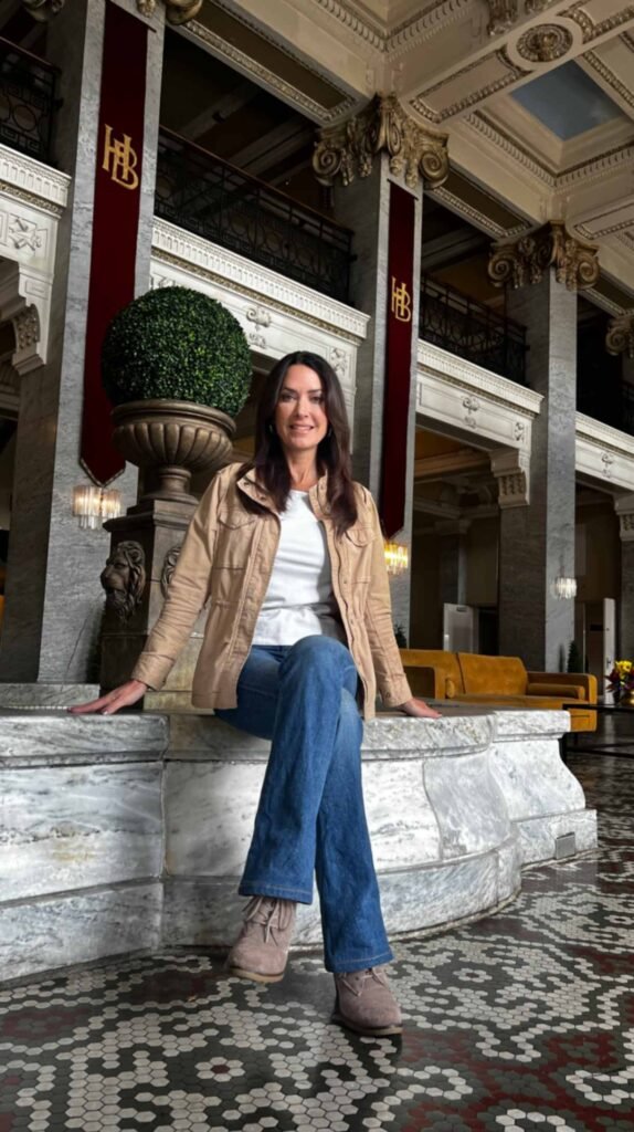 A woman sits on a marble ledge in the historic grand lobby of the Hotel Bentley in Alexandria, Louisiana, showcasing the five-color mosaic tile floor, massive stone columns with ornate gold capitals, and intricate horsehair plaster ceiling molding.