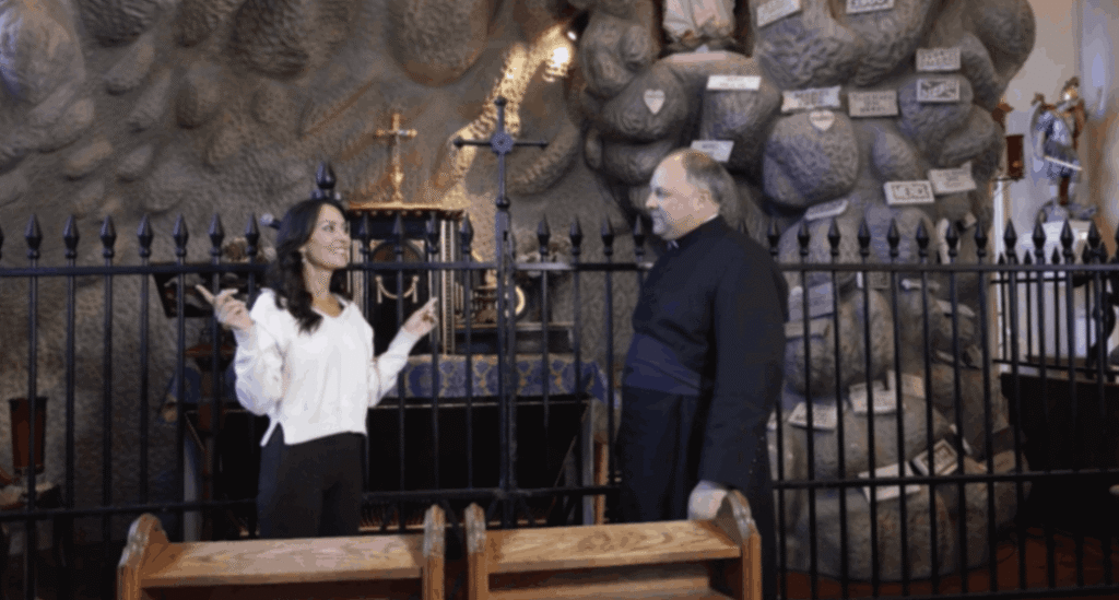 Inside St. Martin de Tours Church, host Karen LeBlanc and Father Jason Vidrine stand before the Lourdes Grotto, a large rock-like formation in the church's transept. The grotto is adorned with numerous white 'Merci' plaques and prayer petitions. A black wrought-iron fence separates the sanctuary area from the grotto, which also features religious statues and a small altar.
