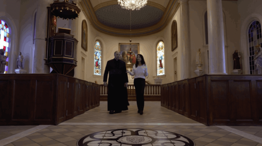 In the center aisle of St. Martin de Tours Church, Father Jason Vidrine and Karen LeBlanc walk toward the altar. The floor features a large circular crest, while dark wooden pews line the sides. Above them hangs a crystal chandelier, and the background shows the ornate sanctuary with stained glass windows and an elevated wooden pulpit to the left.