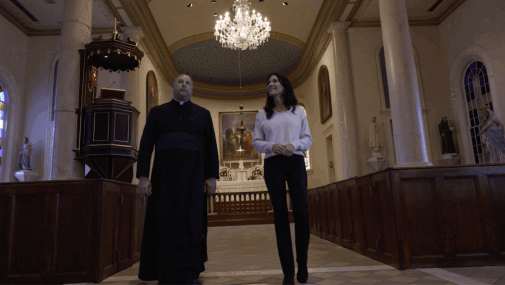 Father Jason Vidrine and Karen LeBlanc walk down the center aisle of St. Martin de Tours Church, looking up toward the high arched ceiling. They are flanked by large Doric columns and dark wooden box pews. In the background, a large crystal chandelier hangs below a blue starlit dome, with the ornate sanctuary and altar visible behind them.