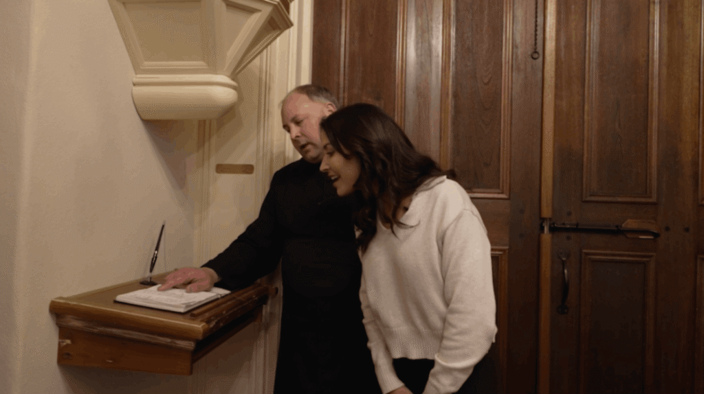 Near a heavy wooden door inside St. Martin de Tours, Father Jason Vidrine points to an entry in a guest registry or historic ledger resting on a wall-mounted wooden podium. Karen LeBlanc leans in to look at the book. A black pen in a holder sits next to the book on the small desk.