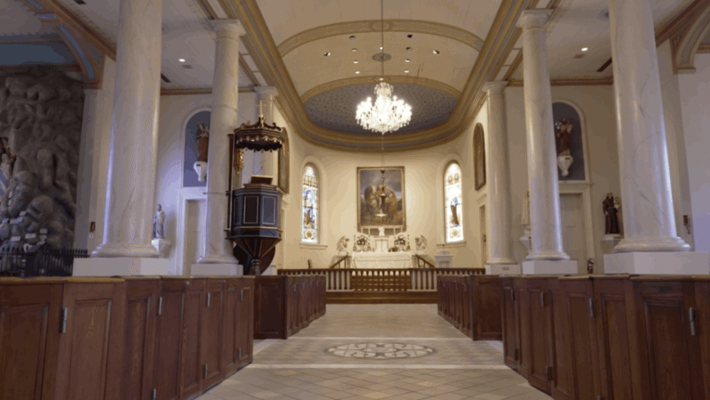 A wide interior view looking down the center aisle of St. Martin de Tours Catholic Church toward the altar. The sanctuary is framed by large white Doric columns and features a prominent crystal chandelier hanging from a blue, starlit dome ceiling. Dark wooden box pews line the foreground, and an elevated wooden pulpit stands to the left of the altar rail.