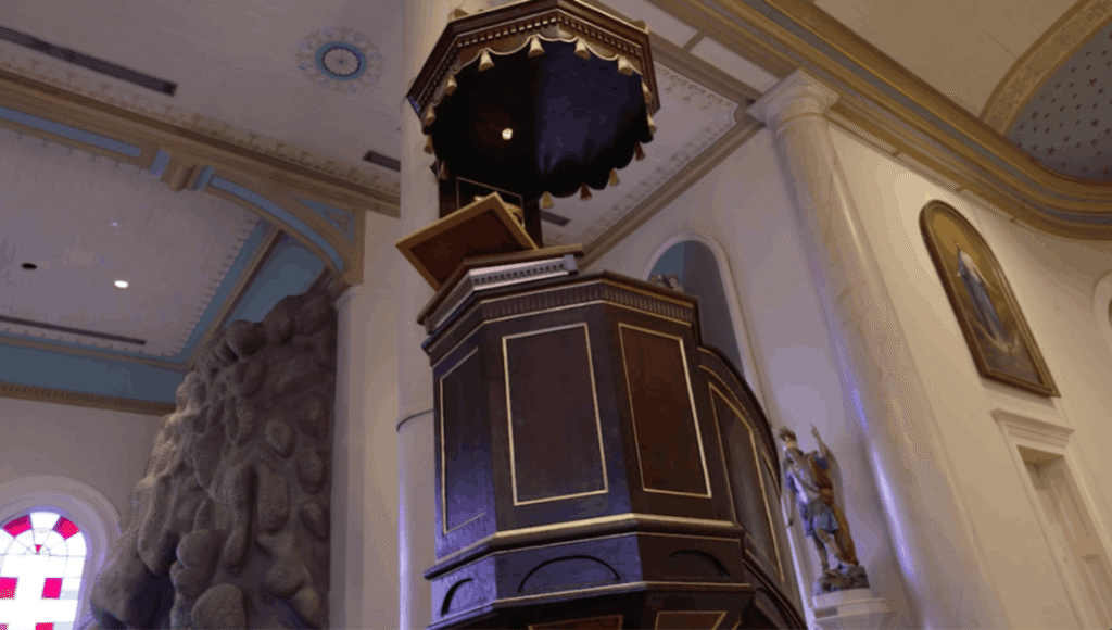 A low-angle view inside St. Martin de Tours Church features an elevated dark wood pulpit with gold trim and a fringed canopy. To the left, the textured gray surface of the Lourdes Grotto rises toward the ceiling near a stained glass window. Large marble-finished columns and a religious painting in an oval frame are visible against the cream-colored walls.