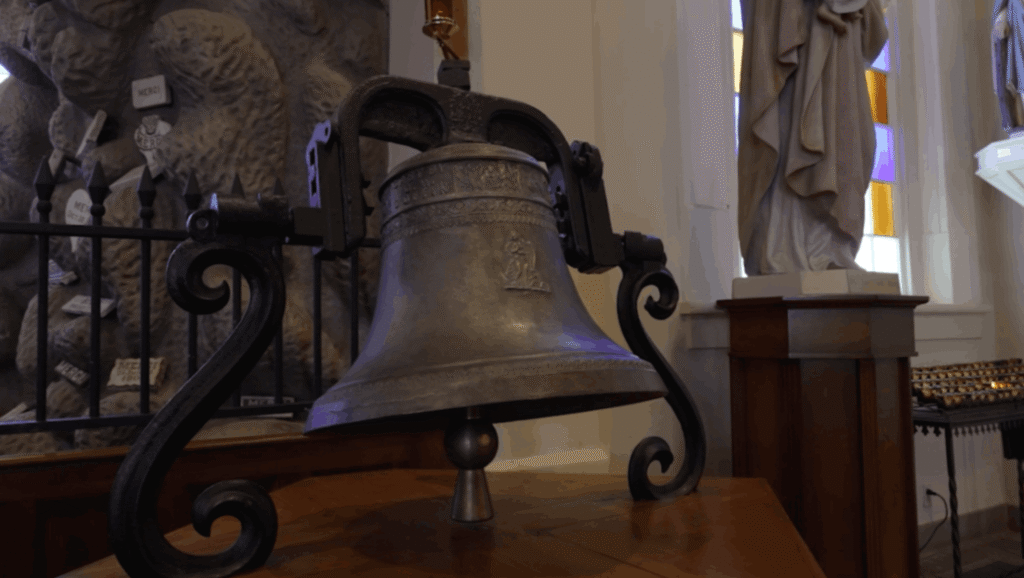 A close-up of an ornate, antique silver-toned bell mounted on a scrolled dark metal stand, resting on a wooden table inside St. Martin de Tours Church. The bell features a relief of a religious figure. In the background, the Lourdes Grotto rock wall and a white religious statue are softly blurred.