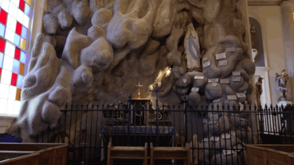 A large, textured rock grotto fills a corner of the St. Martin de Tours Church, next to a tall stained glass window with red and yellow panels. A statue of the Virgin Mary stands in a high niche within the rock, which is covered in small white prayer plaques. A black wrought-iron fence and wooden kneeling rails sit in the foreground before a small altar.