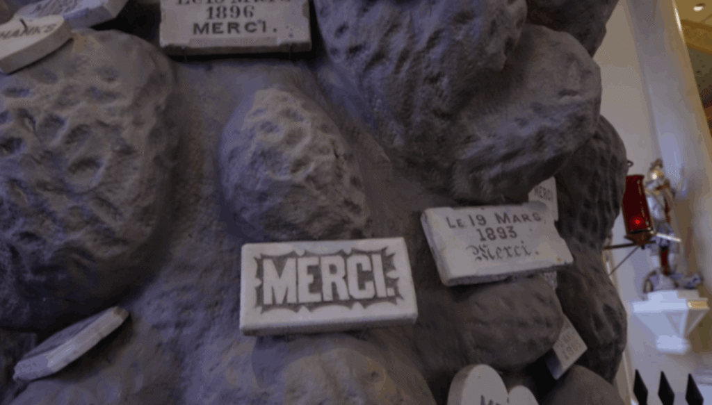 A close-up view of the textured gray surface of the Lourdes Grotto. Several small, white marble 'Merci' plaques of various shapes and sizes are attached to the rock-like wall. One prominent rectangular plaque features the word 'MERCI' in bold black letters, while another nearby is dated 'Le 19 Mars 1893.' A white religious statue is visible in the soft-focus background.