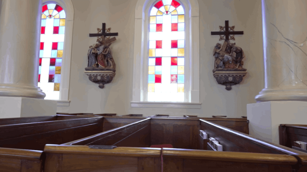 Interior side view of St. Martin de Tours Church showing two stained glass windows with a red, white, and yellow checkered pattern. Between the windows are two ornate, hand-painted 'Stations of the Cross' sculptures mounted on the wall. In the foreground, the tops of high-backed dark wooden box pews are visible between two large, white marble-finished columns.