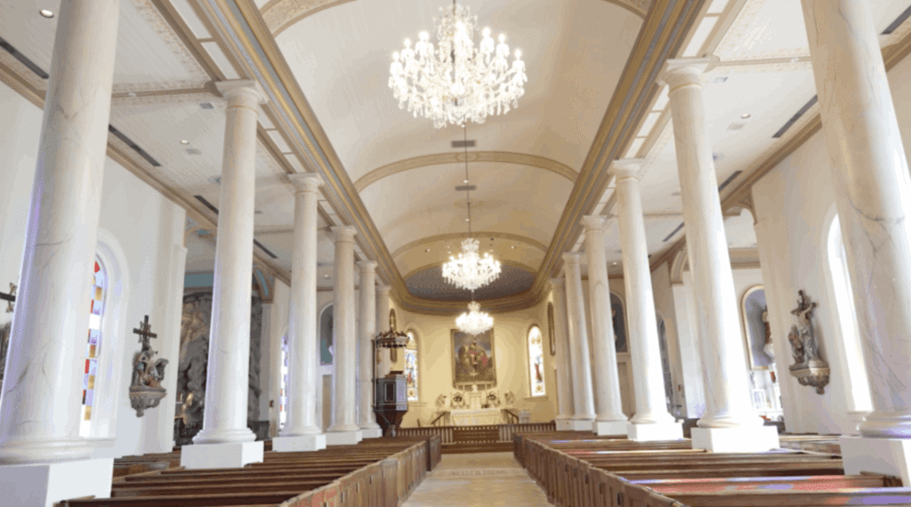 An expansive view down the center aisle of St. Martin de Tours Catholic Church toward the altar. The nave is lined with two rows of tall, white Doric columns supporting a high arched ceiling. Three large crystal chandeliers hang in a line above the aisle. In the background, the sanctuary features a starlit blue dome and an altar painting, while dark wooden pews fill the lower half of the frame.