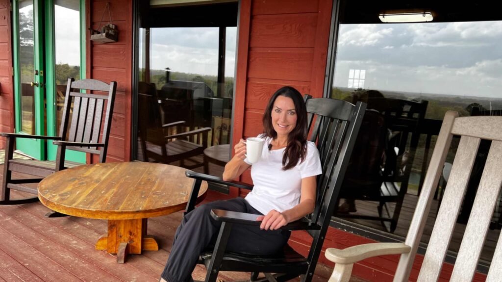 A woman sits on a wooden deck at God's Country RV Park, smiling while holding a white coffee mug. She is seated in a black rocking chair next to a low, round wooden table. The deck features rustic red siding and large glass windows that reflect a scenic overlook of trees and a cloudy sky.