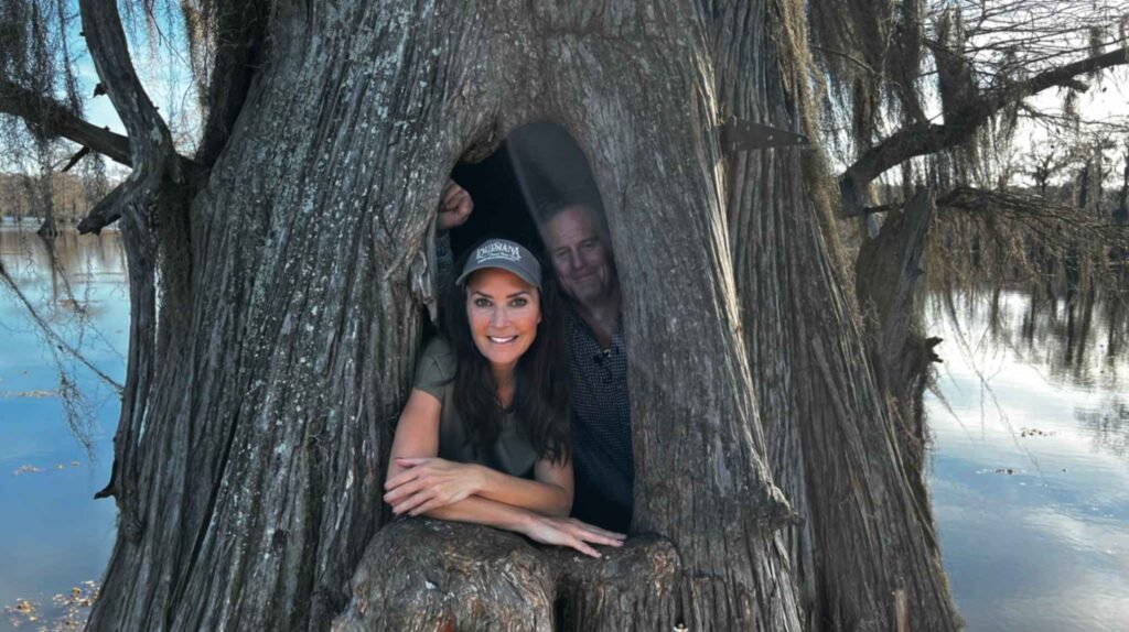 A woman in a "Louisiana" cap and a man smile as they both lean out from a large, natural hollow in the base of a giant bald cypress tree. The massive, textured trunk of the tree dominates the foreground, with the calm, reflective waters of Caddo Lake and distant mossy trees visible in the background.