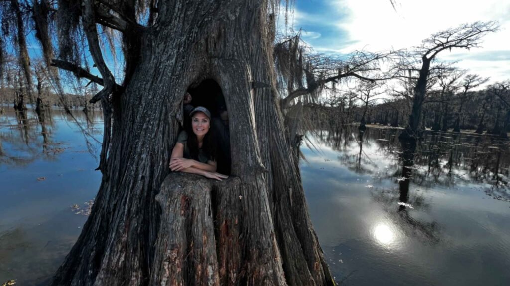 A woman peers out from a large, natural hollow in the massive trunk of an ancient bald cypress tree. The tree stands in the middle of Caddo Lake, with the dark, reflective water and other moss-draped trees stretching out into the distance under a bright, sunny sky.
