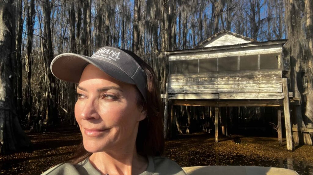 A close-up selfie of a woman wearing a gray "Louisiana" cap, smiling as she looks off-camera. In the background, the weathered wooden Dick and Charlie’s Tea Room stands on stilts over the dark water of Caddo Lake, surrounded by a dense forest of moss-draped cypress trees in the golden afternoon light.