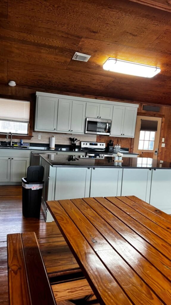 A modern kitchen area inside a rustic lodge at God's Country RV Park. It features light grey cabinetry with black granite countertops, a large kitchen island, and stainless steel appliances including a microwave and stove. A long, polished wood picnic-style dining table sits in the foreground under a wood-paneled ceiling.