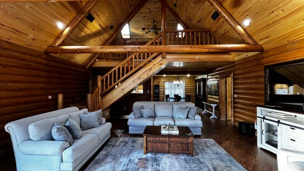 The expansive, open-concept living area of a luxury log cabin at God's Country RV Park. It features two large grey sofas arranged around a dark wood coffee table on a grey area rug. The room has high vaulted ceilings with exposed log beams, a wooden staircase leading to a loft, and log-paneled walls throughout.